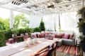 Outdoor patio with white pergola, sectional sofa with pink cushions, striped rug, and dining table surrounded by greenery.