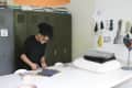 Person cutting fabric on a table in a workspace with lockers, books, and fabric samples on the wall.