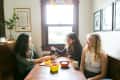 Three women sitting at a wooden table, sharing tea and fruit in a cozy dining nook with framed art on the walls.