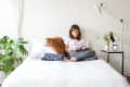 Woman reading on a bed with white bedding, surrounded by plants, a fringed pillow, and a side table with flowers and a lamp.