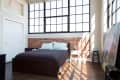Loft bedroom with large windows, exposed brick wall, dark bedspread, and a painting leaning against the wall.