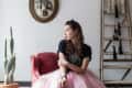 Woman in a pink tulle skirt sitting on a red chair, next to a cactus and a rustic ladder, with a bird artwork on the wall.