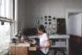 Woman in a white shirt working at a wooden desk in a craft room with tools and supplies on pegboard.