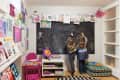 Two children drawing on a large chalkboard in a colorful playroom with books and artwork displayed.