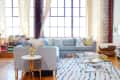 Bright living room with a gray sectional sofa, colorful pillows, glass coffee table, and stacks of books by large windows.