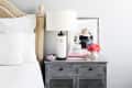 Bedside table with a white lamp, framed photo, pink flower, decorative bicycle, and books next to a white bed.
