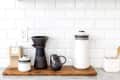 Coffee brewing setup with ceramic dripper, black mug, white carafe, and coffee bag on wooden tray against tiled backsplash.