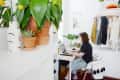 Person working on a laptop at a small desk, surrounded by plants and clothes on hangers in a bright room.