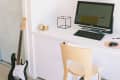 Home office with a wooden desk, computer, chair, wall shelves, and a black electric guitar leaning against the wall.