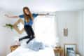 Woman jumping on a bed in a bright room with wooden side tables, plants, and wall decor.