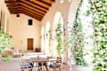 Covered patio with wooden table and chairs, arched openings, climbing plants, and lantern-style wall lights.