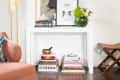 White console table with framed art, books, a green floral vase, and a brass bowl, next to a pink chair and a wooden stool.