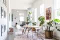 Dining area with round wooden table, clear chairs, large potted plants, and a chandelier, adjacent to a hallway with framed art.