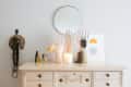 Vanity with decorative hand stands, jewelry, perfume bottles, a lit candle, and a round mirror on a white dresser.