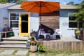 Patio with orange umbrella, yellow door, white chairs, colorful cushions, and potted plants on a wooden deck.