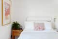 Bed with white linens, red patterned pillow, wooden side table, potted fern, and abstract wall art.