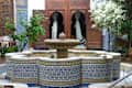 Moroccan courtyard with a central fountain, blue mosaic tiles, wooden doors, and lush greenery.