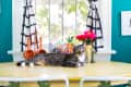 Tabby cat lounging on a yellow table with colorful mugs, pink flowers, and striped curtains in a bright room.