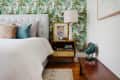Bedroom with tropical leaf wallpaper, tufted headboard, colorful pillows, wooden nightstand, lamp, and vintage fan.