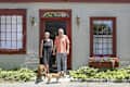 Couple standing with two dogs in front of a brick building with red trim and flower boxes.