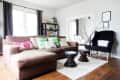 Brown sectional sofa with colorful pillows, black armchair, wooden tables, and bookshelf in a bright living room.