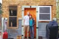 Couple standing on porch of a rustic tiny house with wood and metal siding, red shovel, and "Party On" sign.