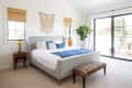 Bright bedroom with a gray bed, blue throw, patterned bench, macramé wall hanging, and large plant by sliding glass doors.