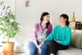 Two people sitting on a gray sofa, laughing, with a colorful rug, potted plant, and wall art in a bright living room.