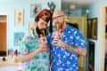 Two men in floral shirts drinking from tiki mugs in a colorful room with eclectic wall art.