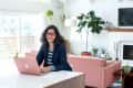 Woman with red glasses using a laptop in a bright living room with a pink sofa and large plant.