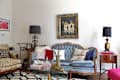 Eclectic living room with a blue tufted sofa, vintage artwork, patterned armchair, and decorative lamps on side tables.