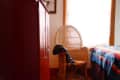 Wicker chair with a black jacket draped over it, next to a bed with a colorful patterned blanket and a red cabinet.