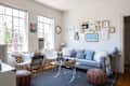 Living room with gray sofa, gallery wall, white chairs, glass coffee table, and brown poufs.