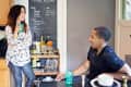Two people chatting in a kitchen with a drink cart, chalkboard, and green glassware.