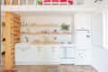 White kitchen with open shelves, farmhouse sink, potted plant, and white appliances under a lofted bed.