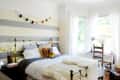 Cozy bedroom with striped walls, brass bed, colorful pillows, and a garland. Bright windows with sheer curtains and a wooden chair.