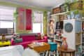 Colorful living room with pink sofa, abstract art, bookshelves, and plants. Geometric rug and eclectic decor complete the space.