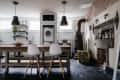 Industrial-style kitchen with a wooden island, white chairs, hanging lights, and a vintage stove next to a fireplace.