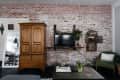 Exposed brick living room with wooden armoire, wall-mounted TV, green armchair, and coffee table with flowers.