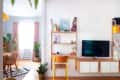 Living room with wall-mounted TV, wooden shelves, yellow chair, and plants, leading to dining area with pink curtains.