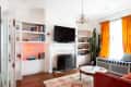 Living room with orange curtains, cactus, neon heart light, bookshelves, and a wall-mounted TV above a fireplace.