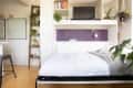 A sleek bedroom with a dark half wall above the bed and a ladder filled with plants in the corner.