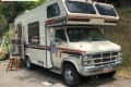 Old GMC motorhome with faded paint and open door, parked on a driveway with a ladder and stool nearby.