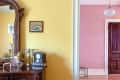 Ornate mirror on yellow wall, vintage clock, globe, and decor on dresser, leading to pink room with wooden door.