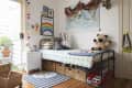 Child's bedroom with a metal bed, colorful bedding, world map, stuffed bear, and storage baskets underneath.