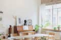 Dining room with a wooden table, chairs, piano, sculptures, framed art, and potted plants by a large window.