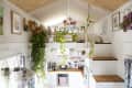 Lofted tiny house interior with plants, wooden stairs, white kitchen, and colorful wall art.