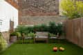 Urban backyard with wooden seating, string lights, lush greenery, and toy trucks on artificial grass.