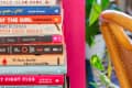 Stack of colorful books on a pink shelf next to a wooden chair and green plant.