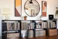 Vinyl records on black shelves with a round mirror, abstract art, potted plants, and a turntable on a wooden floor.
